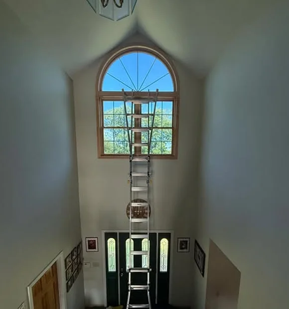 Tall interior window installation scene with a high extension ladder reaching a large arched window above a front entryway