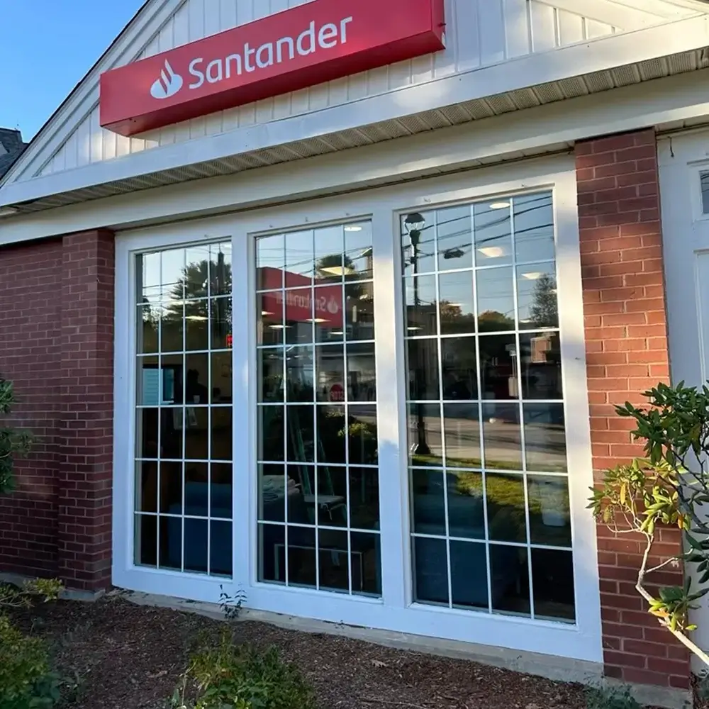 Large front windows of a Santander bank location, reflecting street and sky, with partial window tint applied.