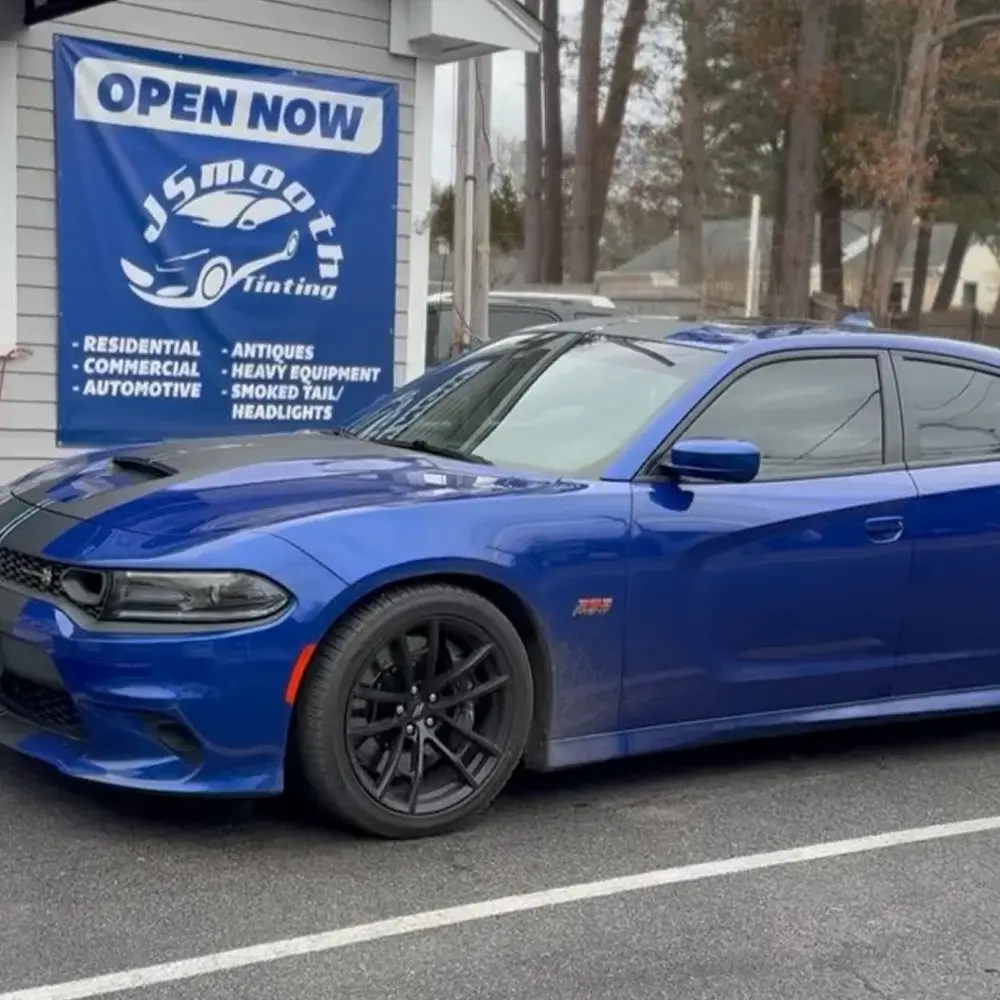 Blue Dodge Charger with tinted windows parked outside a tinting shop with a banner promoting automotive and residential services.