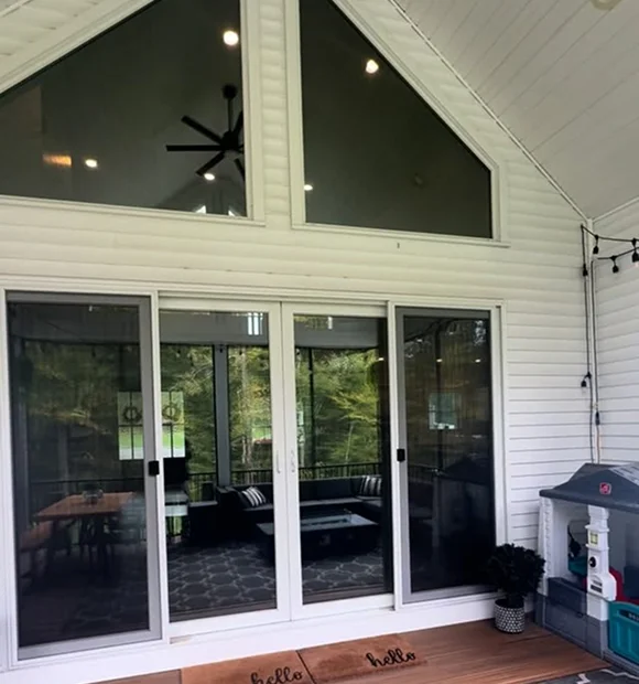 A modern white-paneled sunroom with a high A-frame ceiling and large triangular windows above sliding glass doors. Inside, a cozy seating area with a rug and outdoor furniture is visible through the glass.