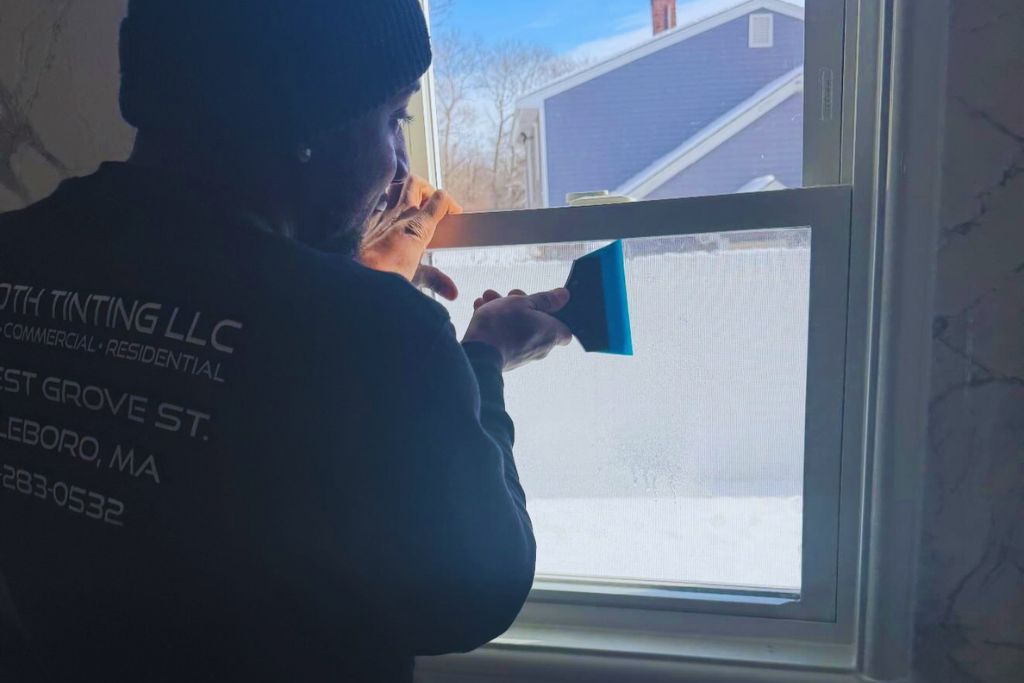 A J Smooth Window Tinting technician uses a blue squeegee to smooth window film onto a residential window, with daylight streaming in and a snow-covered yard visible outside. His shirt displays the company name, location, and contact info, emphasizing professional service.