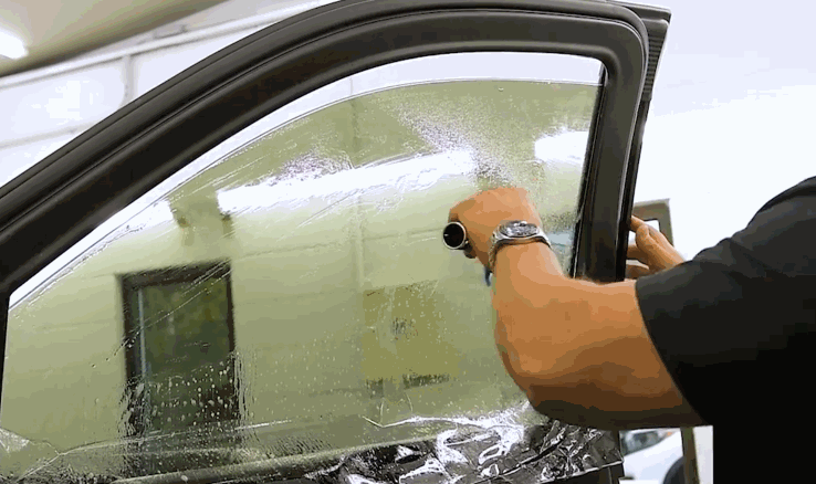 Installer applying window tinting film to a car door window inside a workshop. The glass is covered with moisture as the film is carefully positioned and adjusted.