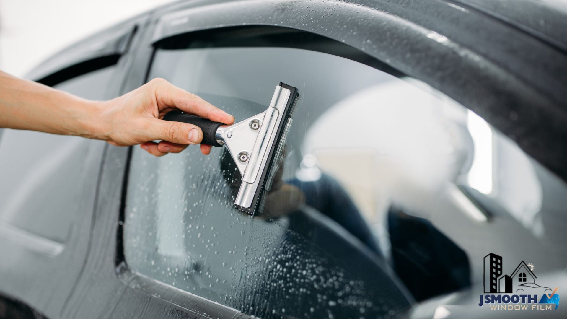 Technician using a squeegee to smooth window tinting film onto a car side window. Water droplets on the glass show the wet application method for a clean and bubble free finish.