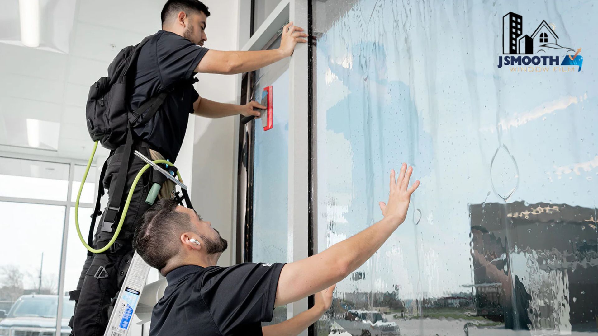 Two technicians install commercial window tint on a large glass window using squeegees and hands to press the film into place. The logo "JSMOOTH WINDOW FILM" appears in the top right corner of the glass.