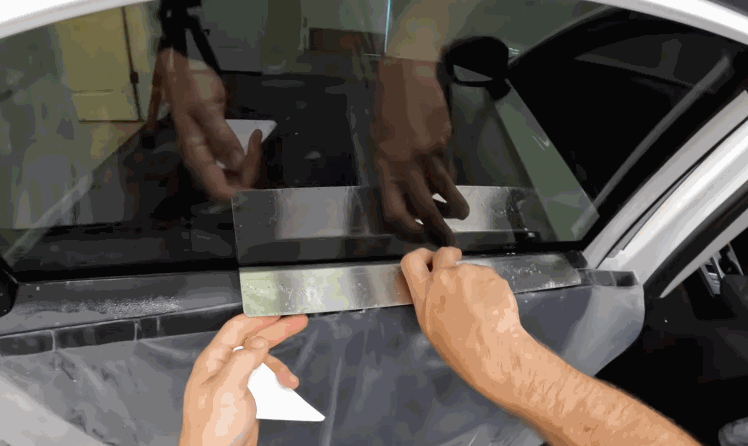 A technician uses a tool to press window film along the lower edge of a car window. The film is being trimmed and secured for a clean ceramic window tint installation.