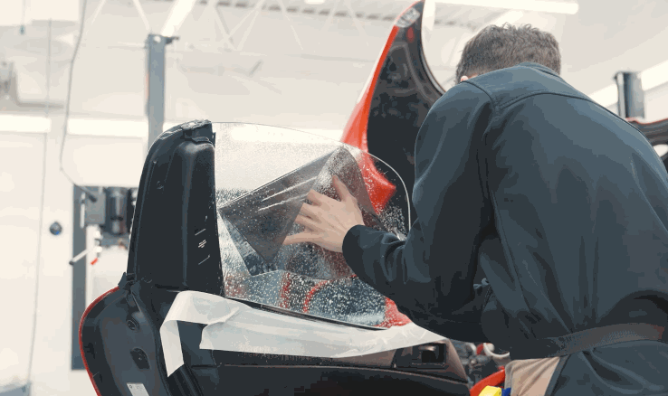 A technician smooths wet window tint onto a car side window from inside the open door. Water droplets cover the glass as the film is fitted into place.