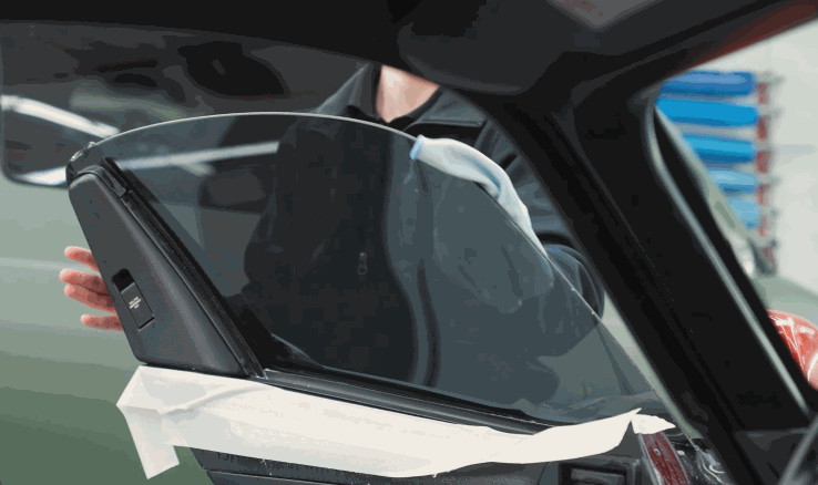 A technician positions window tint on a car side window with tape along the lower edge. The door is open in a bright auto shop.