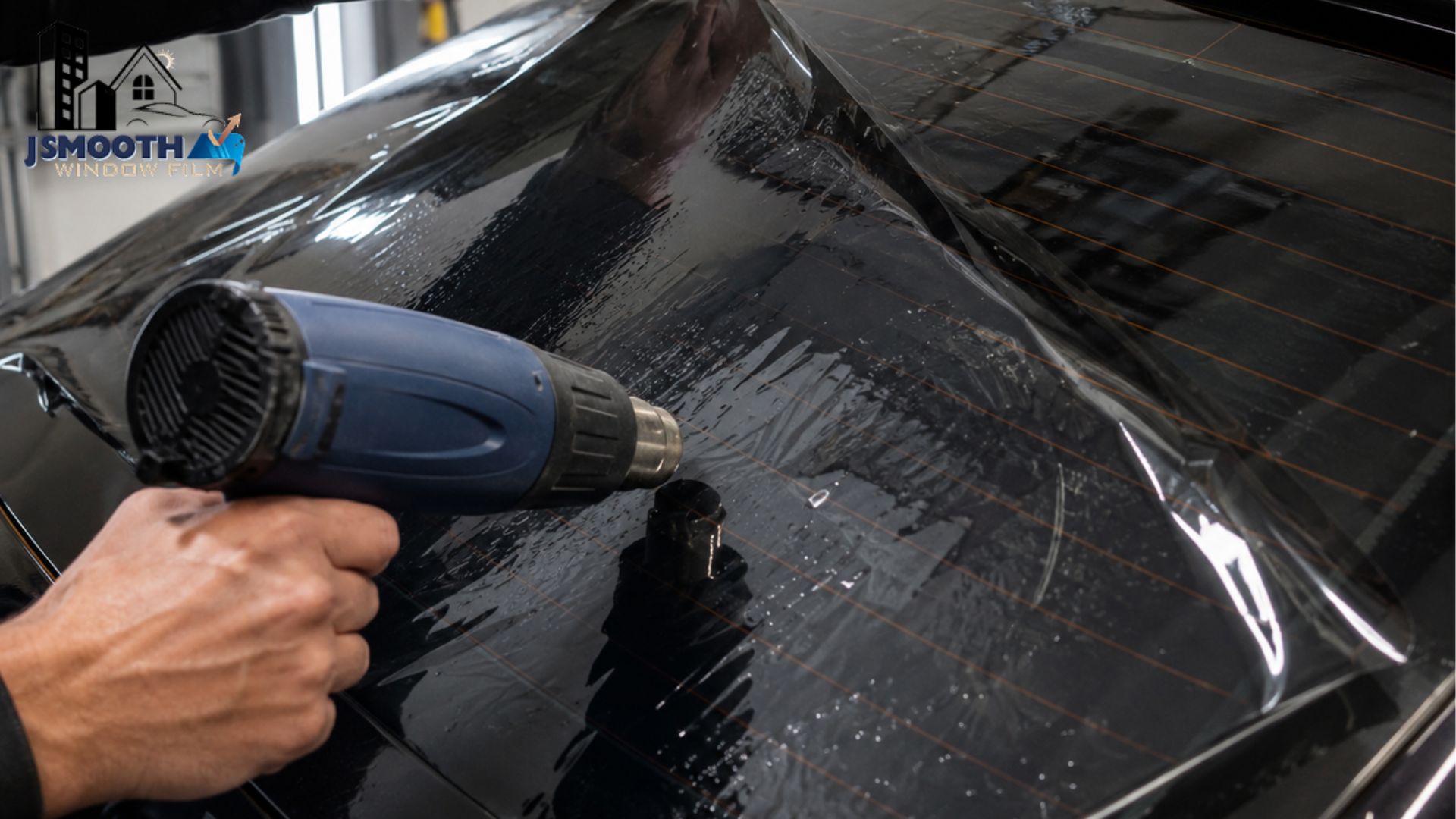 A technician uses a heat gun to shape window tint across the rear glass of a dark vehicle. The text "JSMOOTH WINDOW FILM" appears in the top left corner.