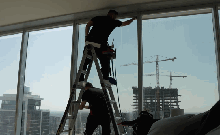 Two installers apply window tint to floor to ceiling apartment windows with cranes visible outside.
